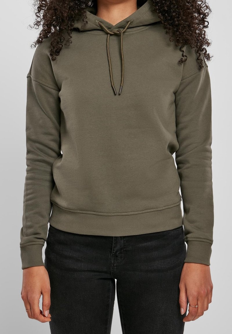Femme portant un sweat à capuche vert olive avec cordons et un jean noir, montrée des épaules aux hanches avec les mains détendues sur les côtés.