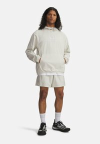 Sudadera con capucha de color beige claro con puños elásticos, bolsillo tipo canguro y logo de "Under Armour". Combinada con pantalones cortos a juego y zapatillas deportivas negras.