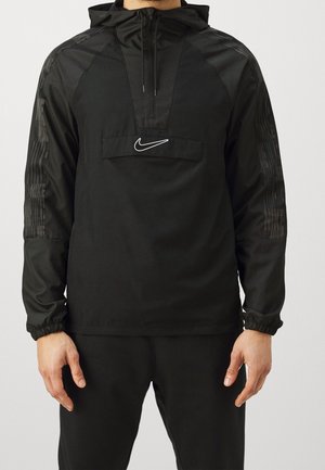 Mężczyzna ubrany w czarną bluzę z kapturem Nike z półzamkiem i logo na piersi, w połączeniu z czarnymi spodniami, stojący na jednolitym tle.