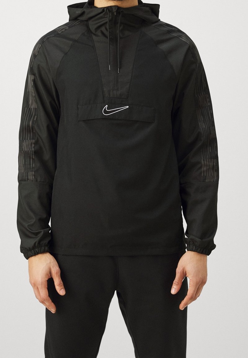 Homme portant un sweat à capuche noir Nike avec une demi-fermeture éclair et un logo sur la poitrine, associé à un pantalon noir, debout devant un fond uni.