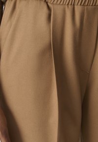 Nahaufnahme von beigen Stoffhosen mit elastischem Bund und Seitentasche, die Textur und Nähdetails zeigen.