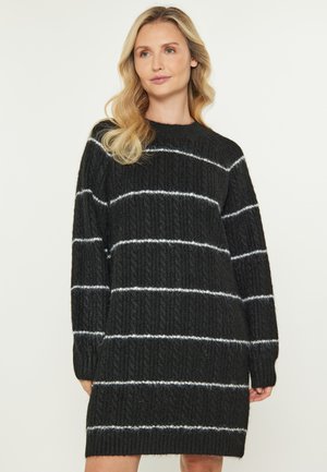 Eine Frau mit blonden, welligen Haaren trägt ein schwarzes Kleid mit Zopfmuster und weißen horizontalen Streifen, während sie vor einem schlichten, hellen Hintergrund steht.