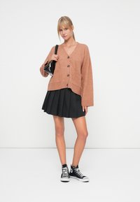 Rustinvärd cardigan med framfickor, runda knappar, avslappnad passform och långa ärmar, kombinerad med en plisserad svart kjol och svarta högklackade sneakers.