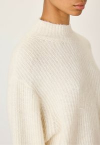 Pullover boje kreme s visokim, rebrastim izrezom, koji ima mekan, teksturiran pletenac i opušteni kroj. Suptilne varijacije u predivu dodaju dubinu.