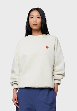 Cremefarbener Sweatshirt aus weichem Material mit rundem Ausschnitt, überschnittenen Schultern und kleinem roten Apfellogo auf der Brust. Abgerundet mit blauen Hosen.