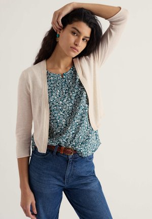 Tânără cu păr lung și închis la culoare, purtând un cardigan bej deschis peste o bluză albastră cu flori și blugi albaștri, pozând cu un braț ridicat.