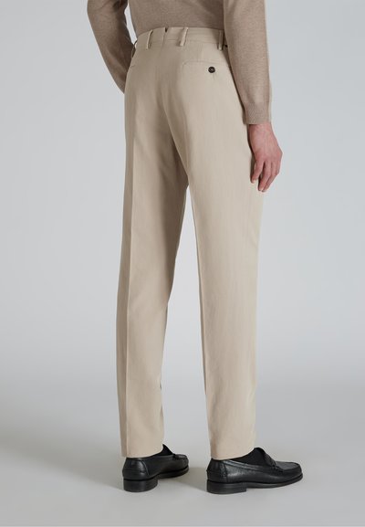 Beżowe spodnie o prostym kroju, z guzikową kieszenią z tyłu, noszone z czarnymi loafersami na neutralnym tle.