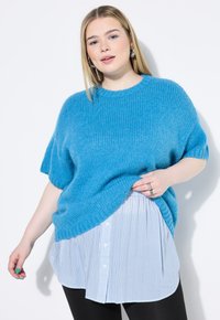 Maglione blu di maglia oversize con maniche corte indossato sopra una camicia bianca e blu a righe, abbinato a leggings neri. Texture liscia.