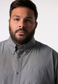 Graues Hemd mit Knopfleiste und strukturiertem Kragen, mit gesticktem "SAILOR" und Zahlen in der linken Brustgegend. Glatte Textur.