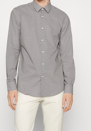 Muž nosící černobílé geometricky vzorované košili s knoflíky a dlouhými rukávy a krémové kalhoty.