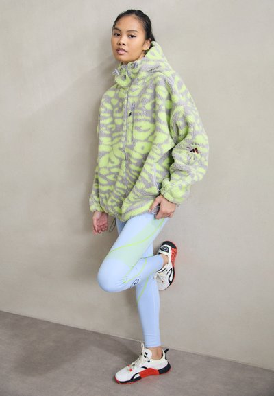 Sweatshirt em felpa oversized em verde limão e padrão de camuflagem cinza, combinado com leggings azul claro com linhas amarelas em contraste e tênis desportivos.