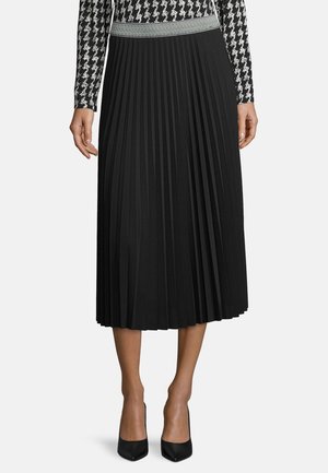 Zwarte geplooide rok met een gestructureerde tailleband, gemaakt van een gladde stof, die tot halverwege de kuit reikt. Draagt zwarte pumps met een spitse neus.