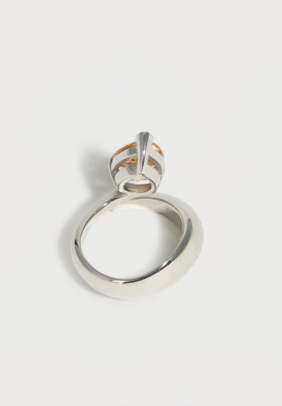 Ασημένιο δαχτυλίδι με παχύ δαχτυλίδι και διακοσμητική, ανοιχτού τύπου στήριξη με χρυσές λεπτομέρειες σε απλό λευκό φόντο.