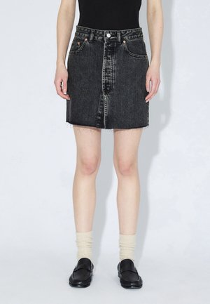 Jupe en denim noir avec un ourlet effiloché, une fermeture éclair à l'avant et deux poches avant. Portée avec un haut noir ajusté, des chaussettes crème et des mocassins noirs.