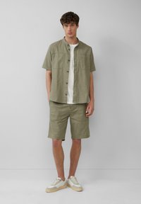 Chemise courte à manches courtes vert olive clair avec un devant à boutons, assortie à un short. Portée avec des baskets blanches avec des accents beiges.