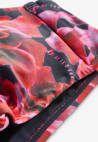 Multicolore stof met grote, abstracte bloemenpatronen in tinten van rood en roze op een zwarte achtergrond, met gestikte randen en merknaam.
