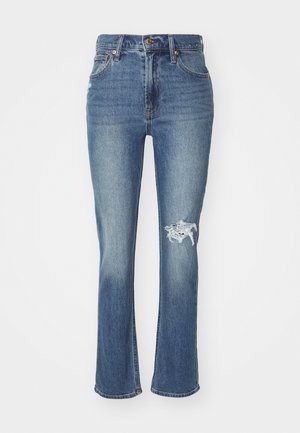 Jean en denim bleu avec un ourlet évasé, taille haute, légère usure au genou gauche et design traditionnel à cinq poches.