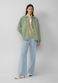 Khaki groene oversized jas met twee grote borstzakken, gecombineerd met een beige top, lichtblauwe wijde jeans en witte sneakers.