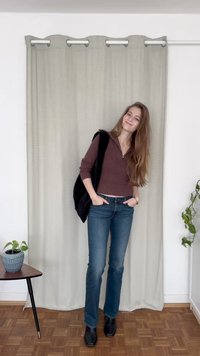 Barna bordázott zipzáras pulóver, sötét mellény, kék farmernadrág és fekete bokacsizma; egy függönynek támaszkodva, egy szobanövény és egy asztal mellett.