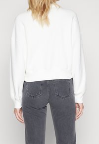 Felpa bianca, corta, con spalle larghe, orlo a coste e maniche lunghe. Abbinata a jeans grigi con tasche posteriori e cuciture.