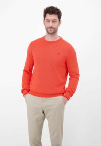 Fel oranje gebreide trui met lange mouwen en geribbelde manchetten. Heeft een ronde hals en een klein logo op de borst. Gecombineerd met beige broek.