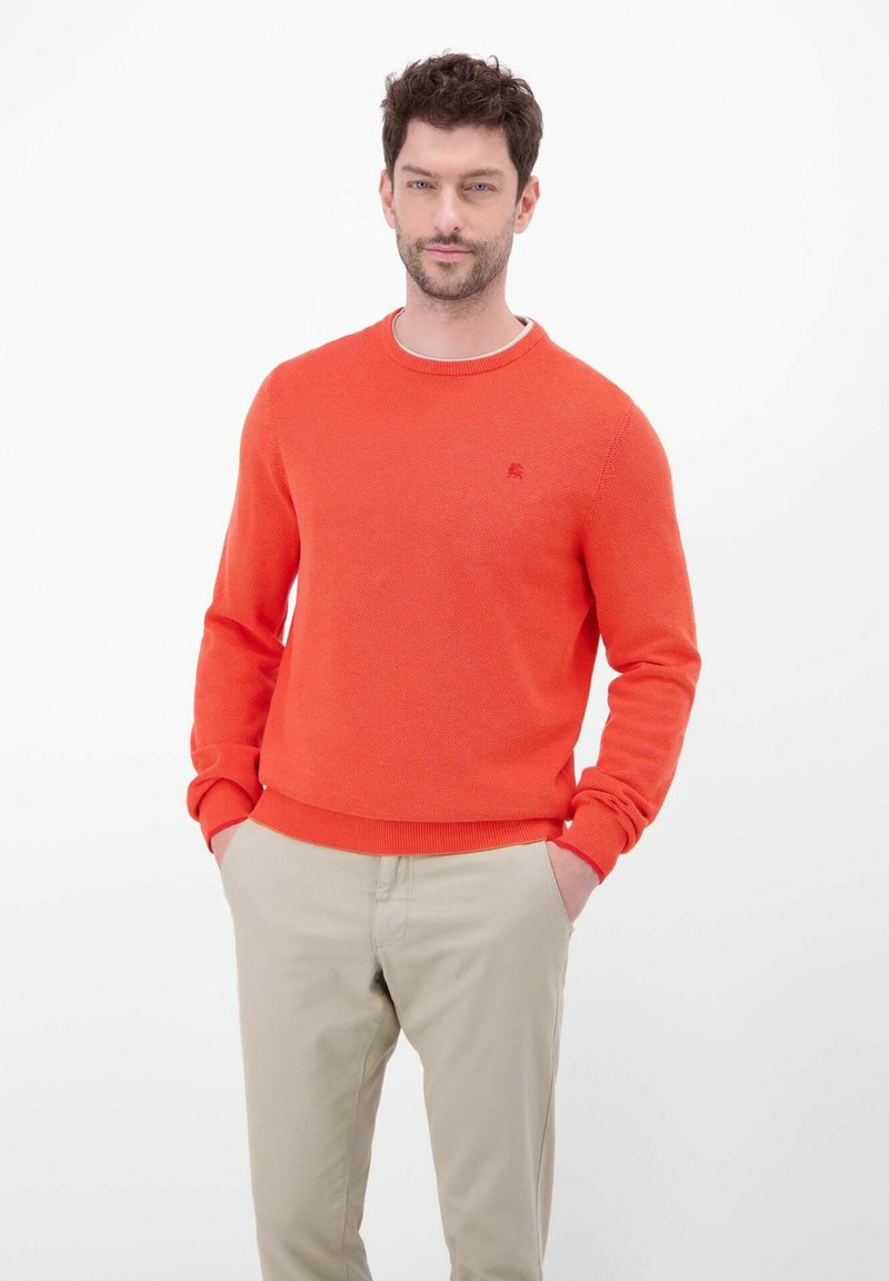 Fel oranje gebreide trui met lange mouwen en geribbelde manchetten. Heeft een ronde hals en een klein logo op de borst. Gecombineerd met beige broek.