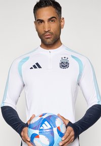 Άντρας με λευκή και ανοιχτό μπλε ποδοσφαιρική φανέλα της Αργεντινής holding μια μπλε και κόκκινη ποδοσφαιρική μπάλα Adidas σε απλό φόντο.