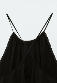 Camisole noire avec des fines bretelles croisées, un décolleté plissé et un tissu texturé agrémenté d'une bordure en forme de scallop sur les bords.
