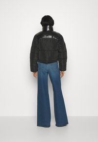 Zwarte cropped pufferjack met een glanzend paneel op de schouders, gecombineerd met wijde blauwe denim jeans. Effen witte achtergrond.