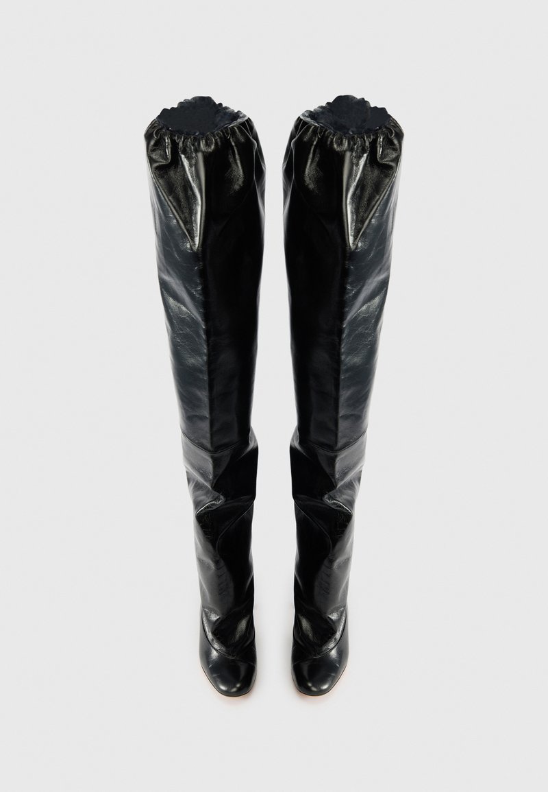 Stivali neri in pelle sopra il ginocchio con una silhouette aderente, bordi superiori arricciati e punte appuntite. Texture liscia con una finitura lucida.
