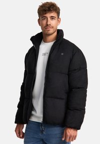 Sort puffersjakke med høj krave, lynlås og struktureret overflade. Båret over en lys sweatshirt og blå jeans.
