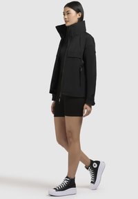 Giacca nera impermeabile con colletto alto, tasche con zip e vestibilità comoda, abbinata a pantaloni corti neri e scarpe sneakers con plateau.