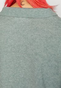 Pull en maille vert clair présentant une texture douce et un col classique, avec un motif subtil et des bords côtelés au niveau de l'encolure.