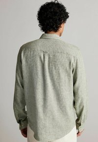 Mężczyzna z kręconymi włosami, ubrany w jasnozieloną, teksturowaną koszulę z długim rękawem, pokazany od tyłu na jednolitym jasnym tle.