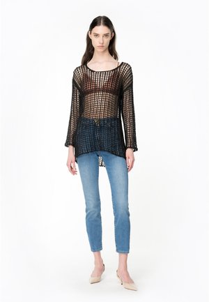Top lungo a maniche lunghe in maglia aperta nera con vestibilità morbida, indossato sopra un top beige, abbinato a jeans azzurri a vita alta e scarpe beige.