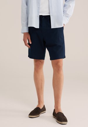 Egy férfi világoskék ingben, fehér pólóban, tengerészkék zsinóros rövidnadrágban és sötétbarna bebújós espadrill cipőben áll semleges háttér előtt.