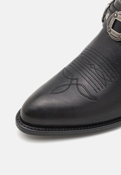 Botín de cuero negro hasta el tobillo con punta afilada, costuras decorativas y detalle de hebilla metálica. Textura suave con un brillo sutil.