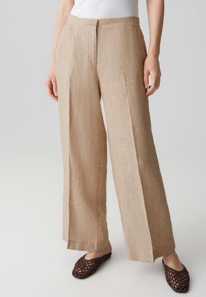 Beige weit geschnittene Leinenhose mit vorderem Knopf und Reißverschluss, getragen mit braunen gewebten Slippern und einem weißen Top.