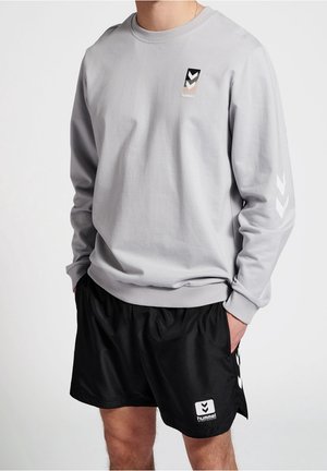 Hombre vestido con un sudadera de manga larga gris claro con un logo en blanco y negro y pantalones cortos deportivos negros con un logo a juego, con las manos en los bolsillos.