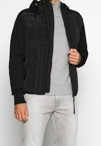 Chaqueta negra con capucha, con una parte delantera acolchada y mangas negras lisas. Cuenta con cierre frontal de cremallera, puños acanalados y bolsillos laterales, llevada sobre una camiseta gris.