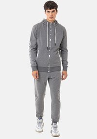 Grauer Zip-Hoodie mit weißen Kordeln und bedruckten Details, kombiniert mit passenden Jogginghosen. Verfügt über Taschen und gerippte Manschetten.