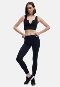 Czarny sportowy crop top z zamkiem z przodu, w zestawie z czarnymi legginsami o wysokim stanie. Buty są czarne z białymi podeszwami, na jednolitym tle.