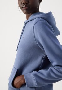Sweatshirt bleu clair avec capuche, poche avant et poignets côtelés. Présente un patch logo sur la poitrine et un tissu doux et texturé.