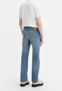 Persona con jeans rectos de color azul claro, una camisa blanca de manga corta y zapatos de ante beige, de pie frente a un fondo blanco.