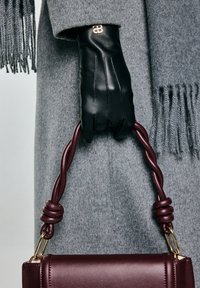 Černé kožené rukavice drží zkroucenou rukojeť bordó kabelky, která je přehozená přes šedý vlněný kabát s třásněmi. Zlaté detaily na rukavicích.