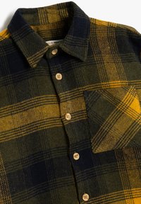 Camisa de franela con un patrón de cuadros en verde, marino y amarillo. Botones de madera destacados y un único bolsillo en el pecho. Textura suave.