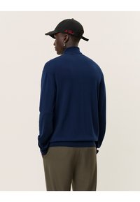 Marineblauer Pullover mit hohem Kragen, langen Ärmeln und geripptem Saum, von hinten gezeigt. Getragen mit olivgrünen Hosen und einer schwarzen Mütze.