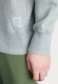 Sudadera gris claro con puños acanalados, que presenta una tela texturizada y un pequeño parche de logo blanco en la manga; combinada con pantalones verde oliva.