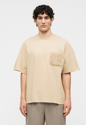 Νέος άνδρας με κατσαρά μαλλιά που φοράει μια μπεζ φαρδιά μπλούζα με υφασμένη τετράγωνη τσέπη, στέκεται μπροστά σε απλό φόντο.