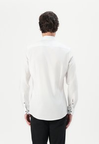 Chemise blanche à manches longues avec un ourlet incurvé, ornée de boutons noirs aux poignets et fabriquée dans un tissu léger et doux.